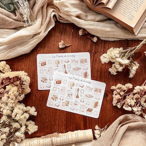 May include: Two white rectangular stickers with a repeating pattern of brown illustrations, including books, globes, and butterflies. The text "Los Pipitas de Guladys" is printed on each sticker. The stickers are on a wooden surface with dried flowers and a book.