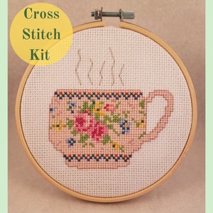 Pode incluir: Kit de ponto cruz com uma xícara de chá floral com vapor saindo do topo. A xícara de chá é rosa com um padrão floral e uma borda amarela e azul. O kit está em um aro de bordado de madeira.