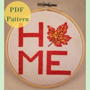 May include: A cross-stitch pattern for a home decor piece featuring the word "HOME" in red with an autumn-coloured maple leaf in the centre. The pattern is displayed in a wooden embroidery hoop.