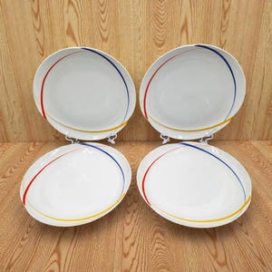 May include: Four white ceramic plates with a colorful design. Each plate has a red, blue, and yellow curved line around the rim. The plates are displayed on white stands against a light brown wooden background.