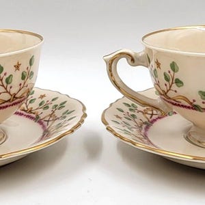 Peut inclure: Deux tasses à thé et soucoupes crème. Chaque tasse et soucoupe a un bord doré et est décorée de feuilles vertes, de volutes dorées et d'une bande violette. Les tasses ont des anses ornées.