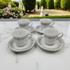 May include: Four white teacups and saucers with a floral design in pink and blue. The rims of the cups and saucers have a silver trim. The teacups are arranged on a white marble surface.