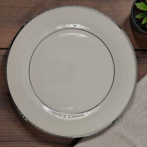 May include: A white ceramic plate with a silver rim and a black inner line. The plate is on a wooden surface, with a small potted plant and a folded linen napkin nearby. The plate is round and has a simple, elegant design.