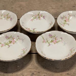 May include: Five white porcelain bowls with scalloped edges and gold trim. Each bowl is decorated with a floral pattern in pink, purple, and green. The bowls are arranged on a wooden surface.