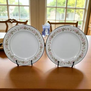 May include: Two white ceramic plates with a floral design border featuring pink flowers and green leaves. The plates have a black trim and are displayed on black metal stands. The plates are on a wooden table.