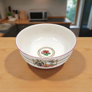 May include: A white ceramic bowl with a decorative fruit and floral design. The bowl has a red rim and a gold and white border with a fruit and floral pattern. The bowl is sitting on a wooden surface.