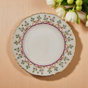 Pode incluir: Um prato de cerâmica branco com uma borda dourada ondulada. O prato apresenta uma borda decorativa de folhas verdes, fitas douradas e pequenas bagas roxas. Um anel roxo e dourado circunda o centro.