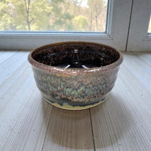 May include: A handmade ceramic bowl with a dark brown interior and a textured exterior. The bowl's rim is a lighter brown, transitioning to shades of blue, green, and yellow. The bowl is sitting on a light-colored wooden surface.