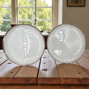 May include: Two white ceramic plates with a gold rim sit on a wooden table. The plates are round and have a simple, elegant design. The background includes a window with a view of trees and a framed picture.