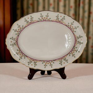 Op de afbeelding: Een ovale serveerschaal met een wit oppervlak, versierd met een decoratieve rand van groene bladeren, paarse accenten en gouden rand. De schaal wordt ondersteund door een donkerhouten standaard, met een klassiek ontwerp.
