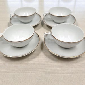 May include: Four white teacups and saucers with gold trim are arranged on a light-colored surface. The cups have a rounded shape with a handle, and the saucers are circular.