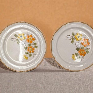 May include: Two off-white ceramic plates with a floral design. The flowers are yellow and orange with green leaves. The plates have a scalloped edge and a brown rim. The plates are on a neutral surface.
