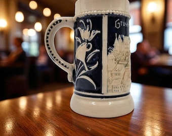 May include: A ceramic beer stein with a white and blue design. The stein features a handle, a floral design, and a relief of a town. The word "Gruss" is written on the side. The stein is sitting on a wooden table.