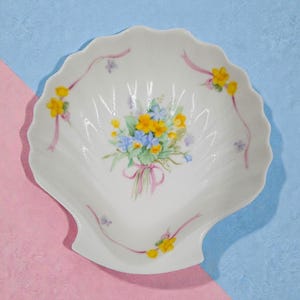 May include: White seashell-shaped dish with a floral bouquet design. The bouquet features yellow and blue flowers tied with a pink ribbon. Pink ribbons and yellow flowers are painted around the rim of the dish.