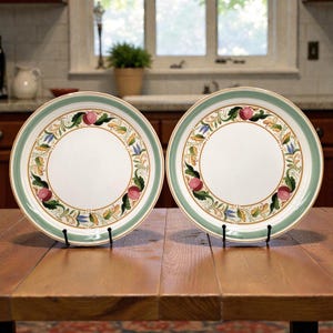 May include: Two white ceramic plates with a green and gold border. Each plate features a decorative ring of pink fruit, green leaves, and blue and yellow floral accents. The plates are displayed on black stands on a wooden table.