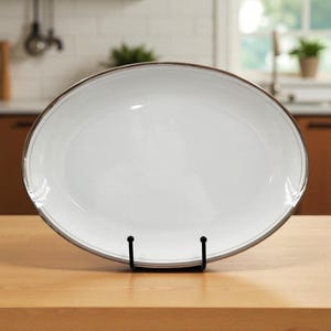 May include: A white oval serving platter with a silver rim, displayed on a black metal stand. The platter is made of ceramic and is set on a wooden surface. The background shows a kitchen setting.