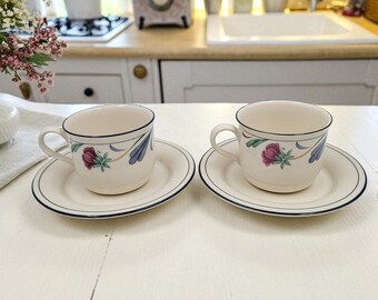 May include: Two matching cream-colored teacups and saucers. Each cup and saucer has a dark blue trim and a floral design with pink and blue accents. The cups are on a white surface.