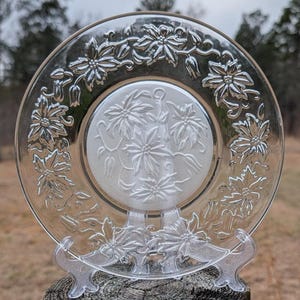May include: Clear glass plate with embossed poinsettia floral design. The plate features a central cluster of flowers and a border of flowers and leaves. The plate is displayed on a clear plastic stand, showcasing the intricate details of the design.