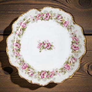 May include: A white porcelain plate with a scalloped edge and a gold rim. The plate is decorated with a floral pattern of pink and white roses. A small cluster of roses is in the center.