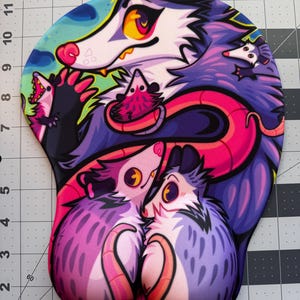 May include: Ergonomic mousepad featuring a colorful illustration of cartoon opossums. The design includes a large opossum with a yellow and pink face, surrounded by smaller opossums in shades of purple, pink, and blue. The mousepad has a wrist rest.