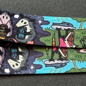 May include: A fabric lanyard featuring a repeating graphic print of opossums. One section shows opossums holding beverage cans against a dark blue background. Another section depicts opossums on a green field, one holding a platter, with flames.