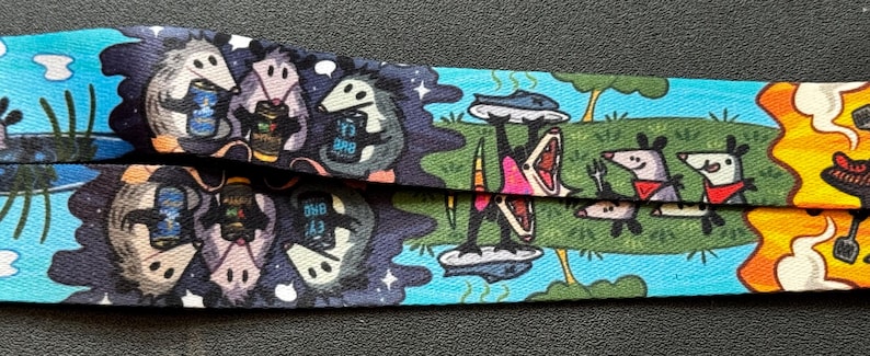 May include: A fabric lanyard featuring a repeating graphic print of opossums. One section shows opossums holding beverage cans against a dark blue background. Another section depicts opossums on a green field, one holding a platter, with flames.