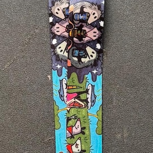 May include: A fabric lanyard featuring a colourful, repeating cartoon print of opossums in various settings, including camping, driving, and playing. The strap is finished with a silver-toned metal lobster clasp at the bottom for attaching items.
