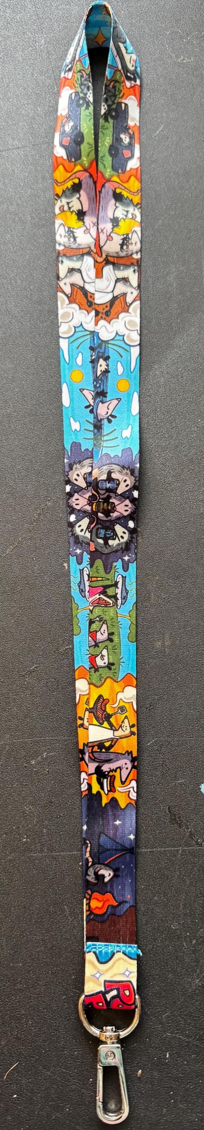 May include: A fabric lanyard featuring a colourful, repeating cartoon print of opossums in various settings, including camping, driving, and playing. The strap is finished with a silver-toned metal lobster clasp at the bottom for attaching items.