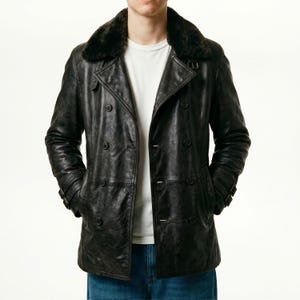 Può includere: Un cappotto doppiopetto in pelle nera con colletto in ecopelliccia. Il cappotto ha diversi bottoni, tasche e un dettaglio con cintura sulle maniche. Sotto si vede una maglietta bianca.