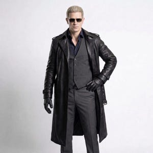 Pode incluir: Um sobretudo de couro preto com colete e calças a condizer. O casaco tem um design longo e fluido com cinto e luvas. A pessoa está a usar óculos de sol e uma camisa azul escura.