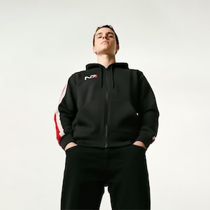 Puede incluir: Sudadera negra con cremallera y detalles en blanco y rojo. La sudadera tiene una franja blanca con una franja roja en las mangas y las letras "N7" en blanco y rojo en el pecho. La persona lleva pantalones negros.