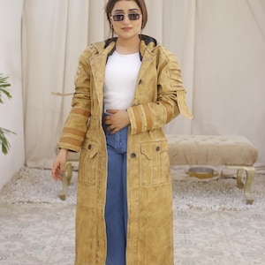 Può includere: Una lunga giacca beige con cappuccio e dettagli in pelle marrone. La giacca ha una chiusura a zip e diverse tasche. La modella indossa jeans blu e scarpe da ginnastica bianche.