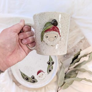 May include: A speckled ceramic mug and matching plate featuring a raised, sculpted gnome character wearing a red hat with a green leaf and dark brown acorn accent. The mug has a side handle, and both pieces have a light cream base with dark speckles.