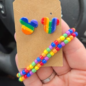 May include: A pair of rainbow Mickey Mouse stud earrings on a brown tag, with a matching beaded bracelet. The earrings and bracelet feature the colors of the rainbow. The bracelet is made of small, colorful beads.