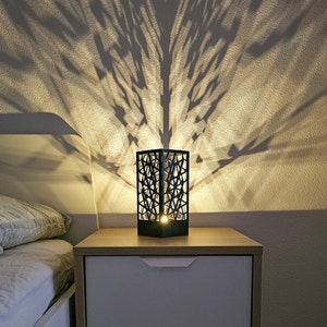 Può includere: Una lampada da tavolo geometrica nera con una lampadina che illumina il design intricato. La lampada è su un comodino in legno con un cassetto bianco.