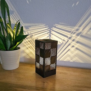 Op de afbeelding: Een zwarte houten lamp met een geometrisch patroon. De lamp is aan en werpt een schaduw op de muur erachter.