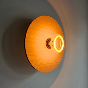 Op de afbeelding: Een ronde, houten wandlamp met een warm, geel licht dat uit een heldere glazen lamp komt.