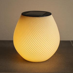 Può includere: Una lanterna da esterno a energia solare con un corpo strutturato color crema e una parte superiore nera. La lanterna ha un design a spirale ed emette una calda luce gialla. Il design è moderno e adatto per l'arredamento di giardini o terrazze.