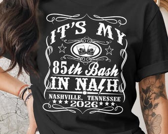 May include: Black t-shirt with white text that reads "IT'S MY 85th Bash IN NASHVILLE, TENNESSEE 2026". The design includes two glasses of whiskey and decorative flourishes.