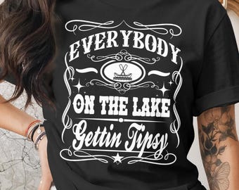 May include: Black t-shirt with white text that reads "EVERYBODY ON THE LAKE Gettin Tipsy." The design includes a boat and oars graphic. The shirt is a casual wear item.