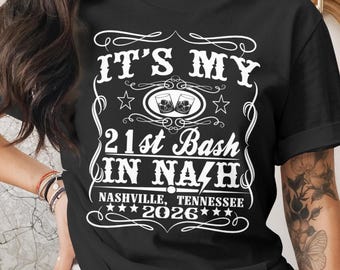 May include: Black t-shirt with white text that reads "IT'S MY 21st Bash IN NASHVILLE, TENNESSEE 2026". The design includes stars and images of two glasses of whiskey.