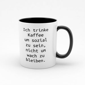 Könnte beinhalten: Weiße Keramiktasse mit schwarzem Rand und Henkel. Die Tasse hat schwarzen Text mit der Aufschrift "Ich trinke Kaffee um sozial zu sein, nicht um wach zu bleiben."