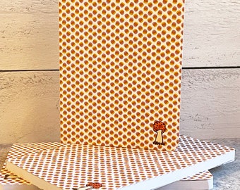 May include: Three notebooks with a repeating pattern of small brown and yellow dots on a cream background. One notebook stands upright, while two others are stacked. A small mushroom illustration is on the bottom right corner of the notebooks.