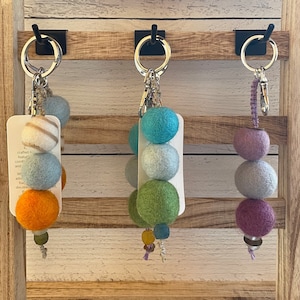 May include: Three keychains with felt balls in different colours. The first keychain has orange, white, and blue felt balls. The second keychain has green, light blue, and turquoise felt balls. The third keychain has purple, light blue, and grey felt balls.