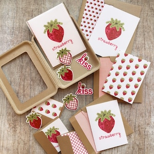 May include: A collection of stationery items featuring a strawberry theme. The image includes greeting cards, stickers, and patterned paper with strawberry illustrations. The cards have a red strawberry graphic and the word "strawberry". The stickers say "kiss".