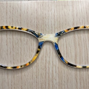 May include: A pair of cat-eye glasses with a silver frame and a colorful pattern featuring blue, yellow, and white. The pattern is reminiscent of a popular science fiction television show.