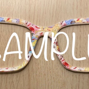 May include: A pair of eyeglasses with a white frame and a colorful print of Disney princesses. The print includes Belle, Cinderella, Snow White, and others. The glasses are labeled "SAMPLE".
