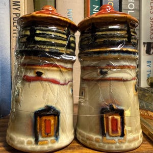 May include: A pair of ceramic salt and pepper shakers. The shakers are shaped like small lighthouses, with a beige base, a dark brown top, and a red accent. The shakers are wrapped in clear plastic.