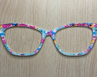 Op de afbeelding: Een paar cat-eye brillen met een blauwe en roze Alice in Wonderland print.