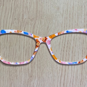 May include: A pair of white cat-eye glasses with a colorful pattern of cartoon characters. The glasses have a white frame with a blue and pink trim.
